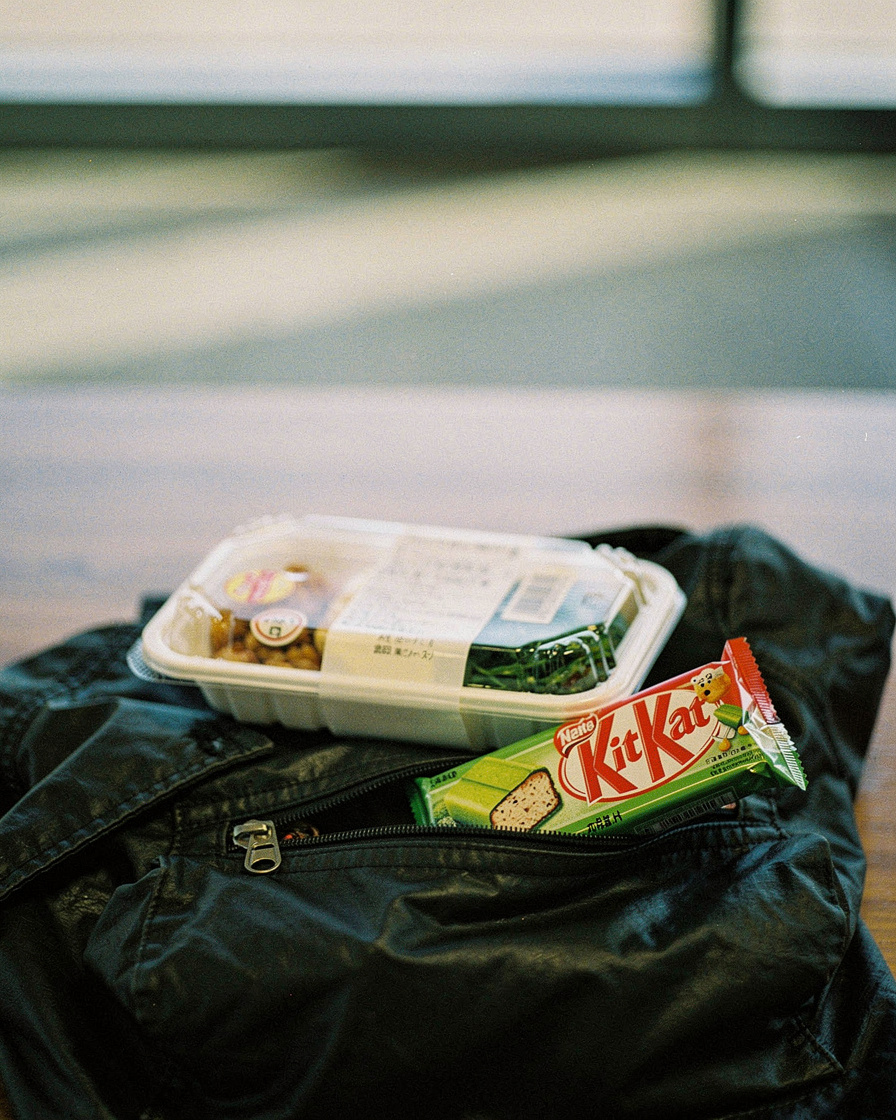 Woman holding a Japanese convenience store bento box at a gate in Kansai Airport, flat overhead terminal light, duffel bag on