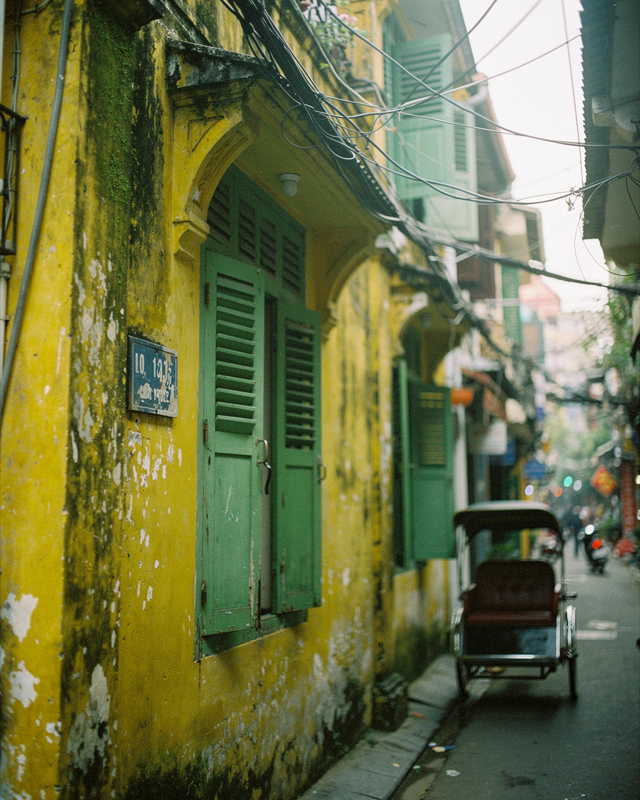 Empty narrow Hanoi Old Quarter street with faded yellow French-colonial buildings, tangled wires overhead, and a parked cyclo
