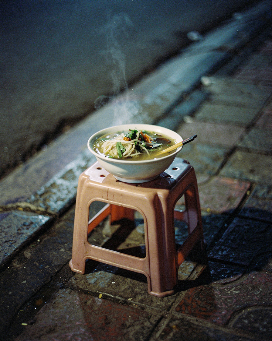 Woman on a low plastic stool eating pho at a street stall on Ta Hien, Hanoi Old Quarter, lit by string lights at night.