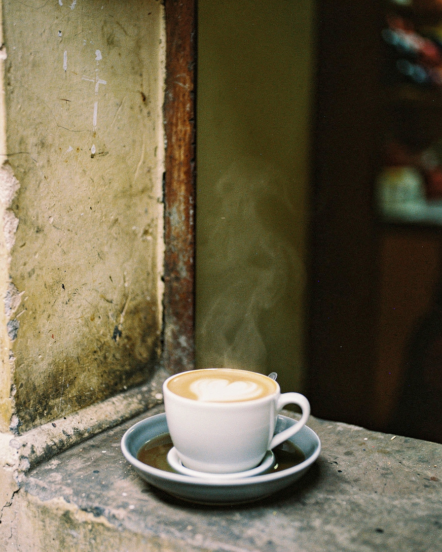 Cup of Vietnamese egg coffee set in a bowl of hot water on a wooden table, soft daylight from a window, Hanoi Old Quarter str