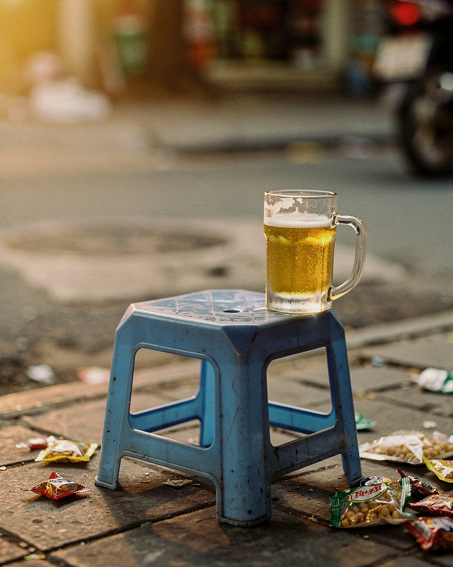 Woman on a low plastic stool at Hanoi's Bia Hoi Junction at dusk, glass of draft beer in hand, street lit by warm bulbs and p