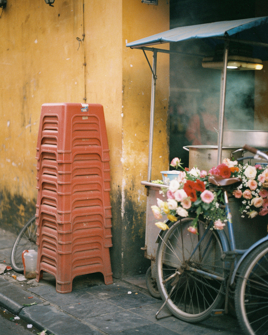 Empty Hanoi Old Quarter street at dawn with a flower-loaded bicycle, stacked plastic stools, and steam rising from a pho cart